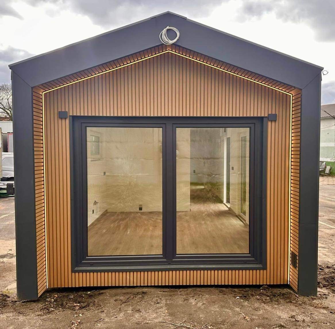 Nowoczesny, modułowy domek z drewnianą elewacją i oknami w Głogowie. Ciemna blacha na dachu, oświetlenie LED podkreśla krawędzie. Widok z zewnątrz, surowy stan.