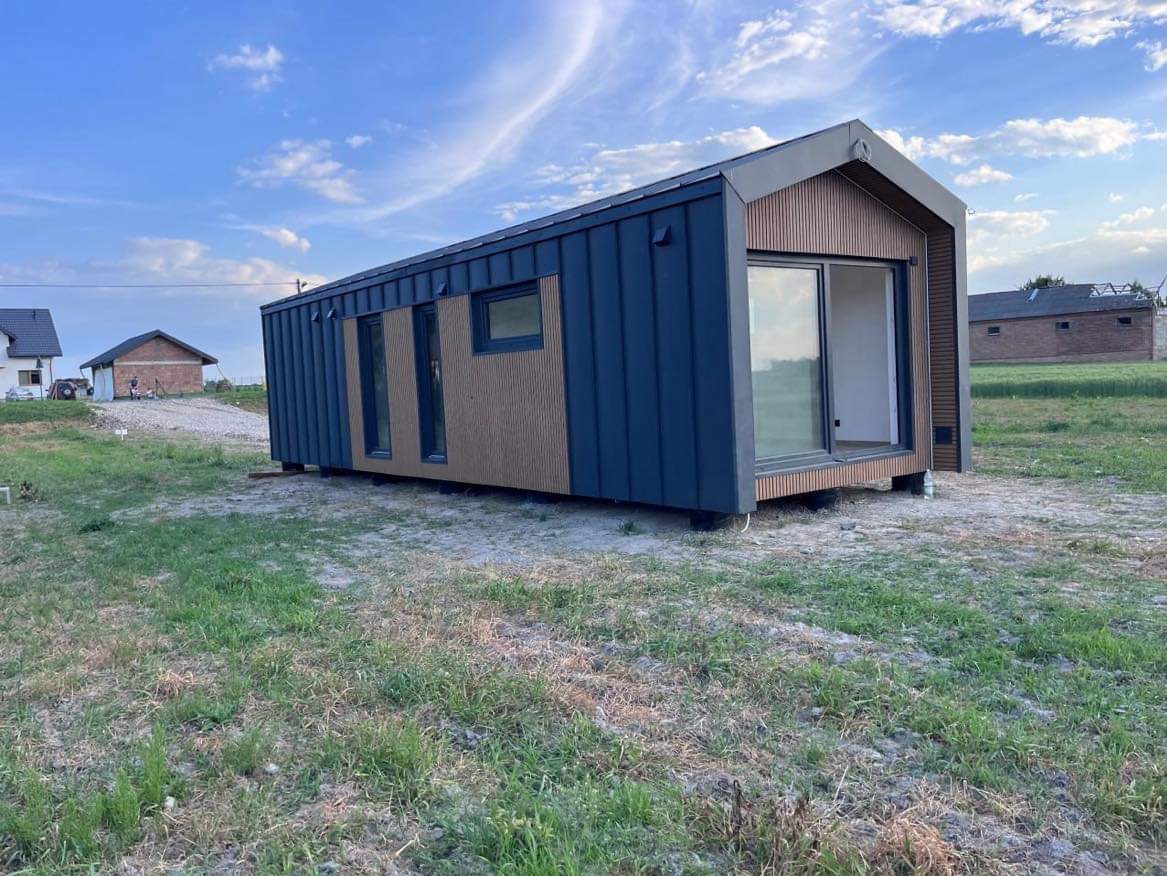 Nowoczesny, modułowy dom z ciemnoniebieską elewacją i drewnianymi akcentami na tle zielonej trawy i błękitnego nieba z chmurami. Widoczne okna i przesuwne drzwi.