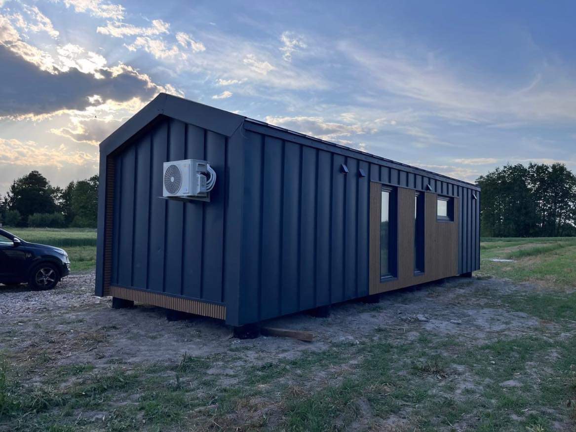 Nowoczesny, mobilny dom w ciemnej okładzinie z klimatyzacją na bocznej ścianie. Widoczny fragment samochodu i zielone otoczenie. Ujęcie z niskiej perspektywy.