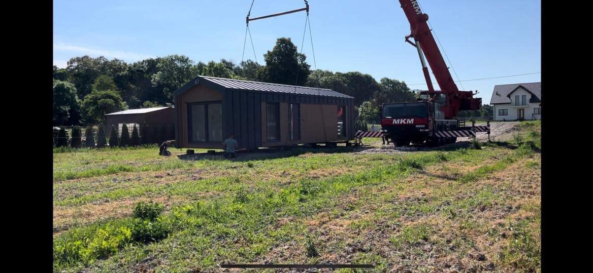 Transport modułowego domu z szarym dachem i drewnianą elewacją za pomocą czerwonego dźwigu na tle zieleni i nieba, widoczny pracownik.