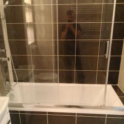 Kamil Rogalski - Wykończona łazienka z czarno-białymi kwadratowymi płytkami, białą wanną z parawanem nawannowym i chromowaną armaturą; widoczne odbicie fotografa w szklanej powierzchni.