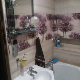 Kamil Rogalski - Wykończona łazienka z białym umywalką, wanną, baterią, lustrem z półką, dozownikiem mydła i ręcznikiem na tle płytek z motywem kwiatowym.