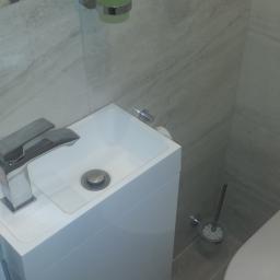 Kamil Rogalski - Kompaktowa, biała umywalka z szafką w nowoczesnej toalecie, uzupełniona o chromowaną baterię, dozownik mydła w płynie i szczotkę do WC na szarej podłodze z płytek.