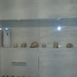 Wykończona toaleta z szarymi płytkami imitującymi marmur, dekoracyjną mozaiką, szafką pod umywalką z szufladą i półką z dekoracjami z muszelek oraz chromowanym przyciskiem spłuczki.