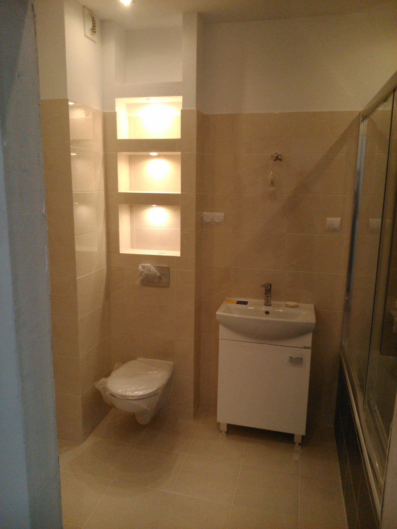 Wykończona łazienka z beżowymi płytkami, podświetlanymi wnękami w ścianie, umywalką z szafką i kabiną prysznicową, toaleta zabezpieczona folią ochronną.