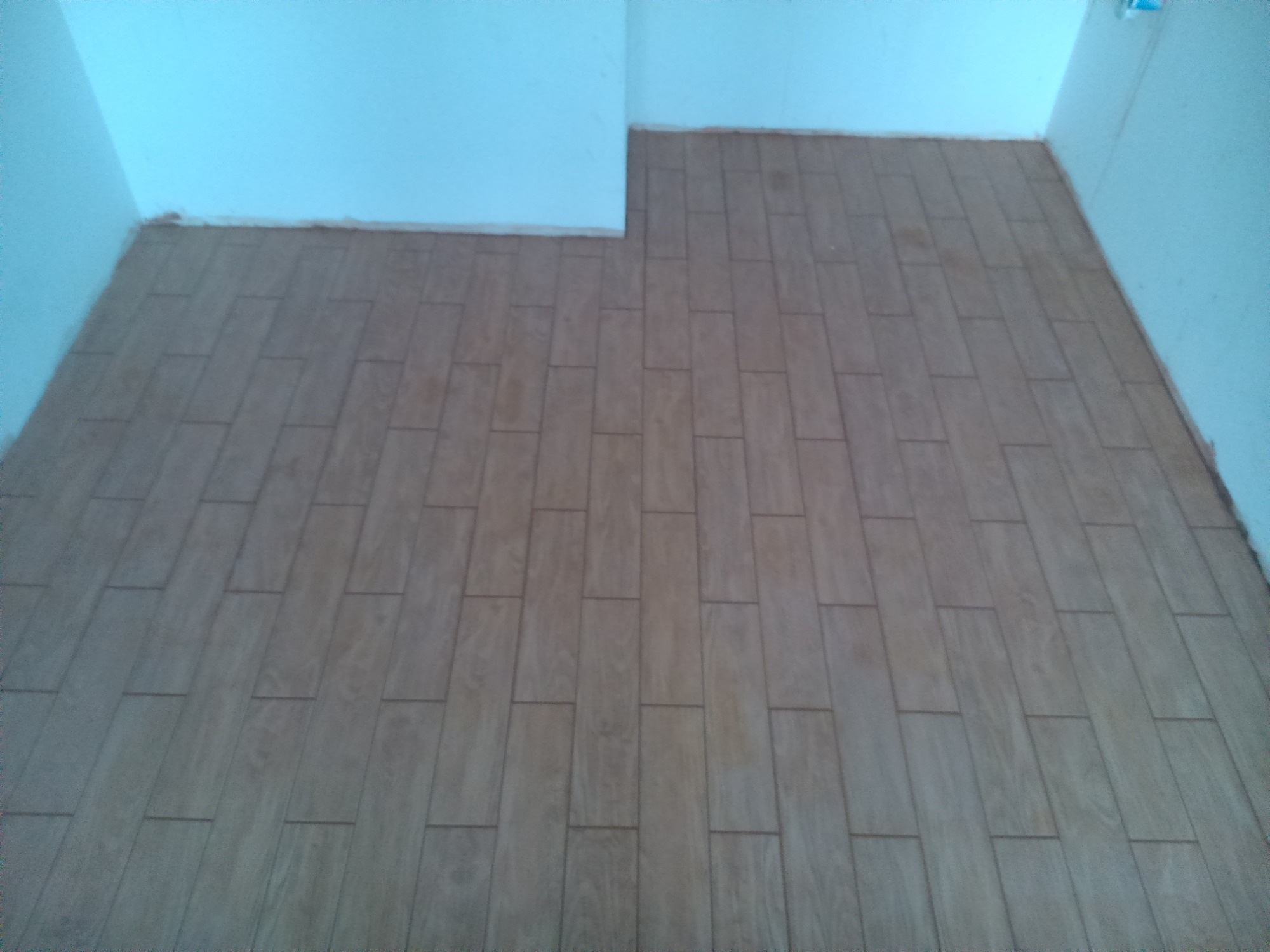 Ułożona podłoga z płytek imitujących drewno w prostokątnym wzorze, widok z góry na fragment pomieszczenia z białymi ścianami, widoczne niedoskonałości przy łączeniu ściany z podłogą.