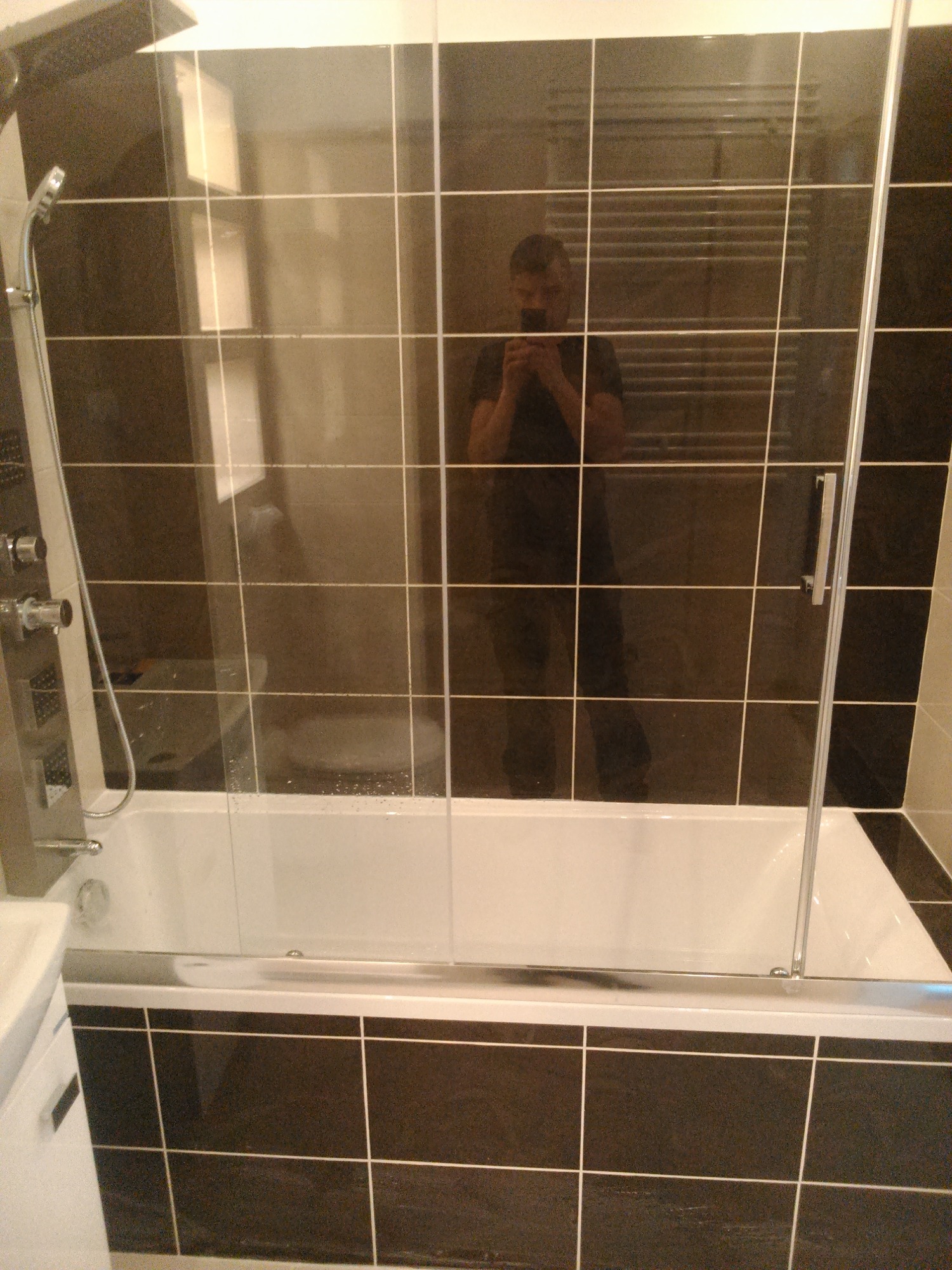 Wykończona łazienka z czarno-białymi kwadratowymi płytkami, białą wanną z parawanem nawannowym i chromowaną armaturą; widoczne odbicie fotografa w szklanej powierzchni.