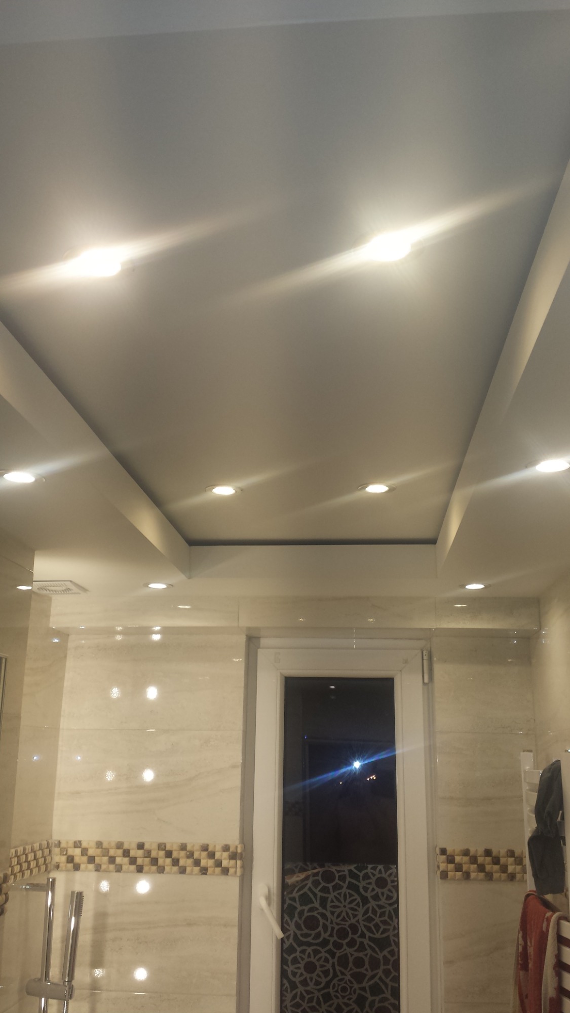 Podwieszany sufit w łazience z oświetleniem punktowym, widoczne płytki na ścianach i okno.