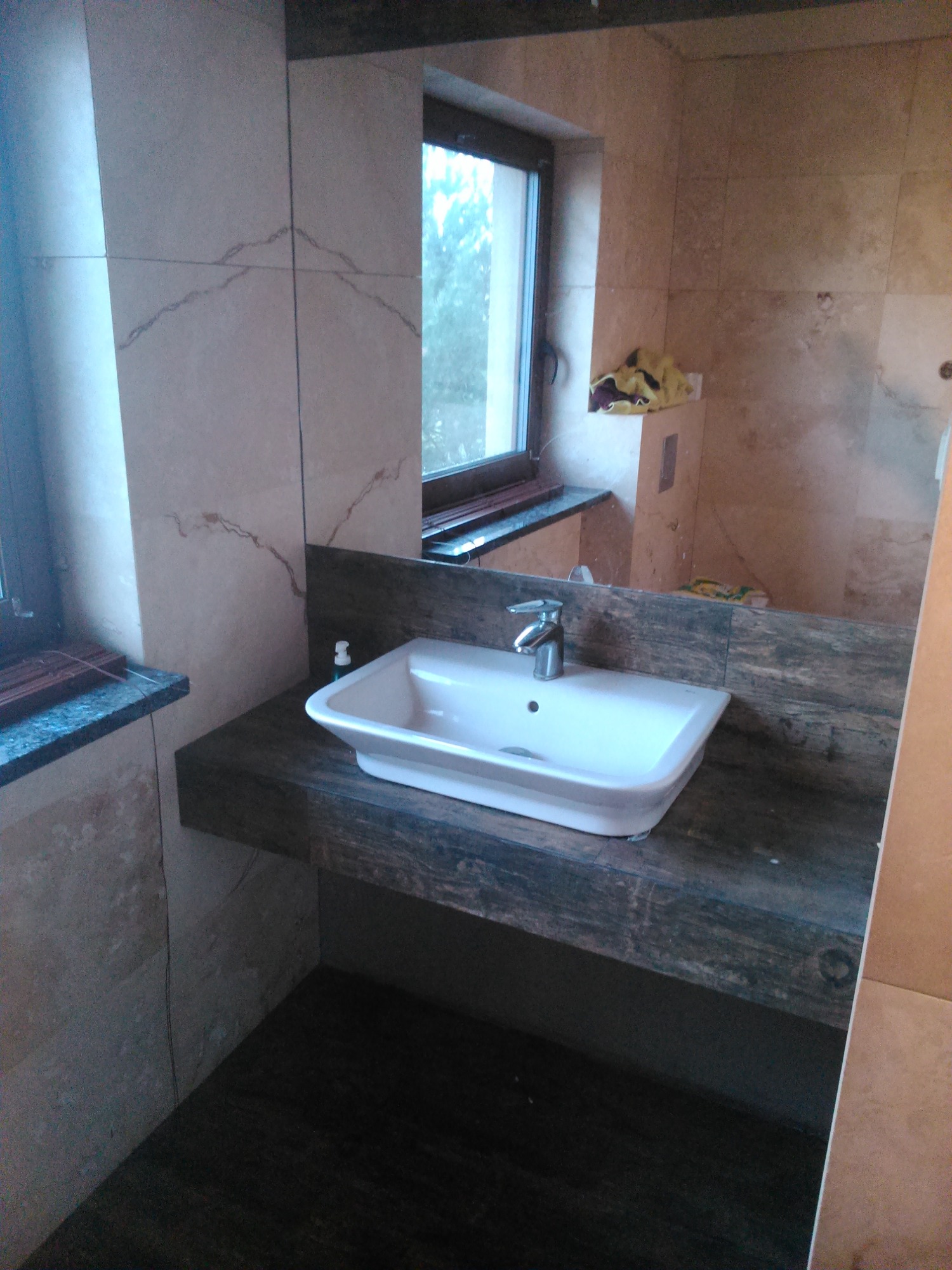 Umywalka nablatowa o kwadratowym kształcie, zamontowana na blacie z ciemnego drewna o wyraźnym usłojeniu, z baterią chromowaną, na tle ściany wyłożonej jasnymi płytkami imitującymi marmur...