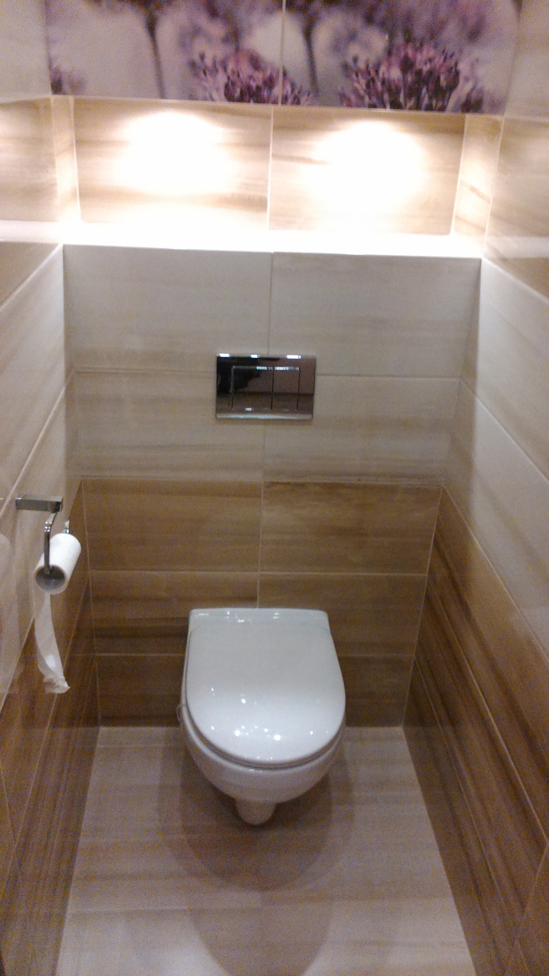 Nowoczesna toaleta w beżowych odcieniach, z podświetleniem LED nad płytkami z motywem kwiatowym i chromowanym przyciskiem spłukującym.