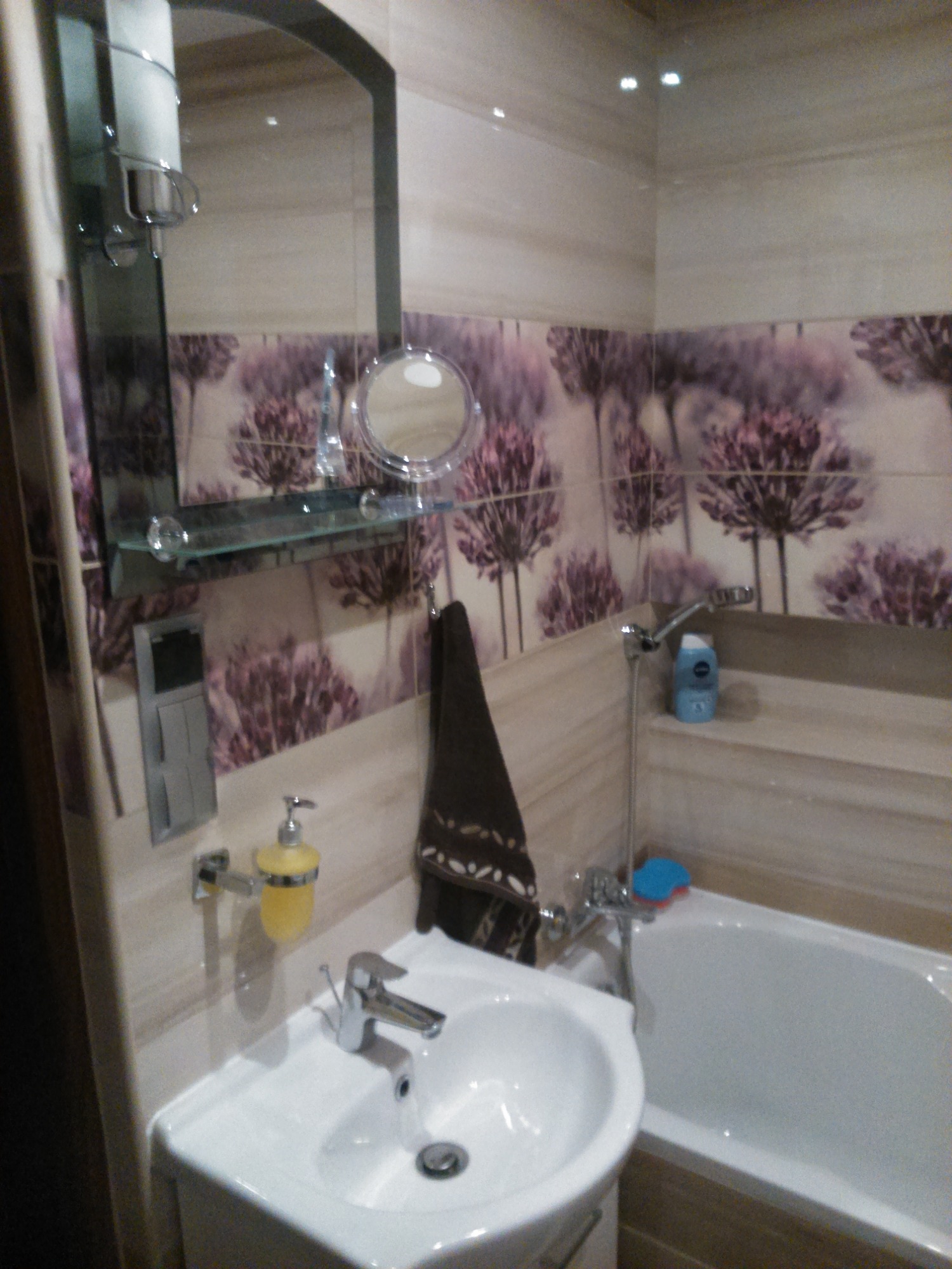 Wykończona łazienka z białym umywalką, wanną, baterią, lustrem z półką, dozownikiem mydła i ręcznikiem na tle płytek z motywem kwiatowym.