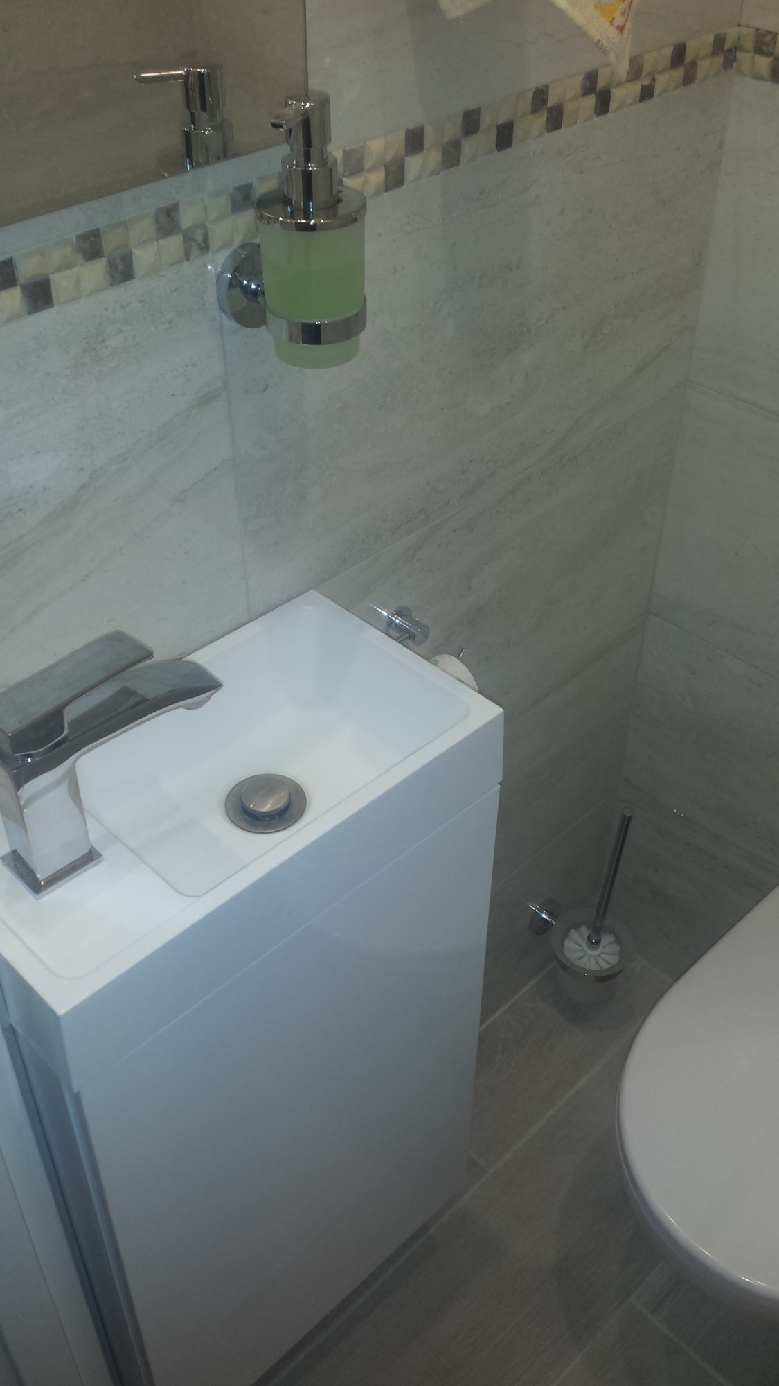 Kompaktowa, biała umywalka z szafką w nowoczesnej toalecie, uzupełniona o chromowaną baterię, dozownik mydła w płynie i szczotkę do WC na szarej podłodze z płytek.