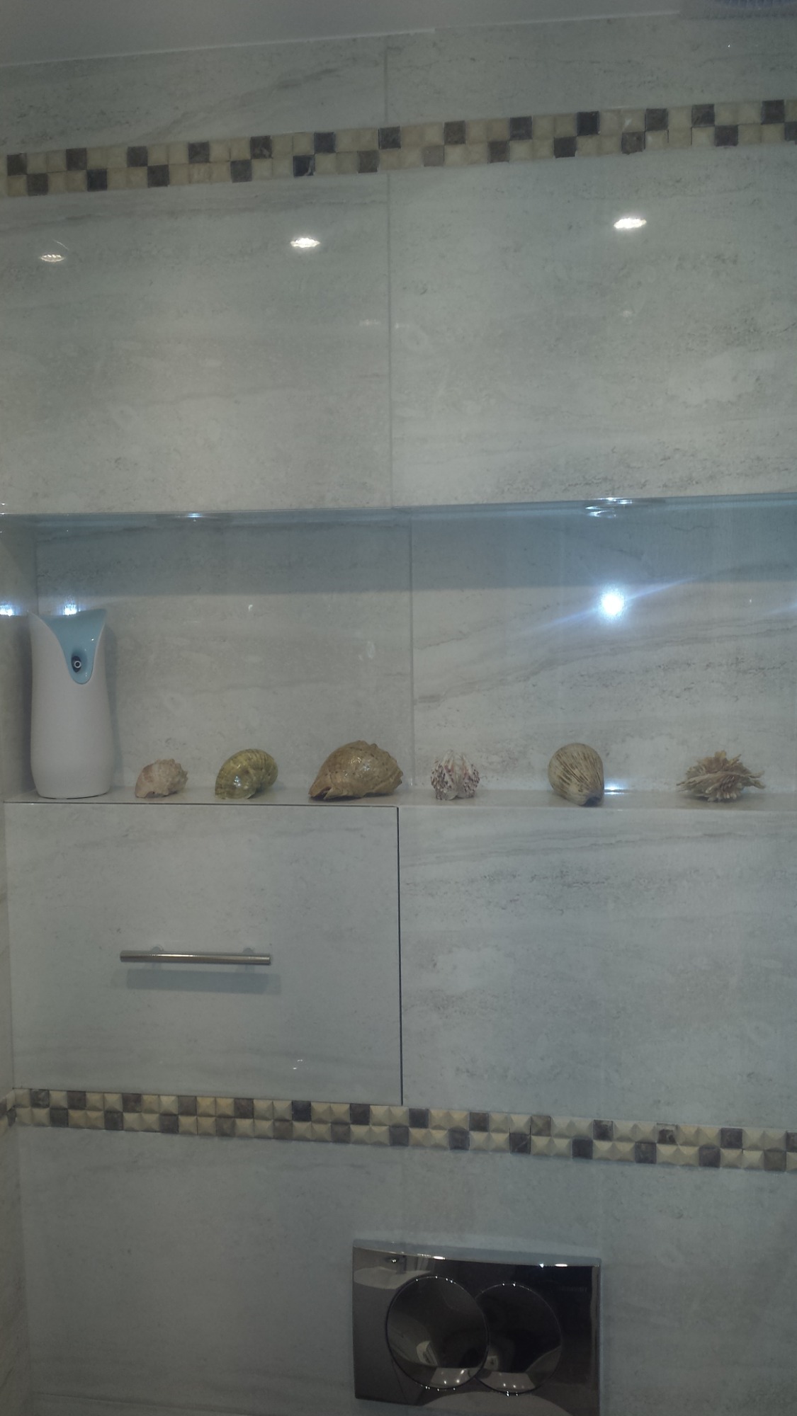 Wykończona toaleta z szarymi płytkami imitującymi marmur, dekoracyjną mozaiką, szafką pod umywalką z szufladą i półką z dekoracjami z muszelek oraz chromowanym przyciskiem spłuczki.