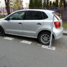 Srebrny samochód osobowy z widocznymi uszkodzeniami karoserii, zaparkowany na chodniku z kostki brukowej, obok stoi biała łata miernicza.