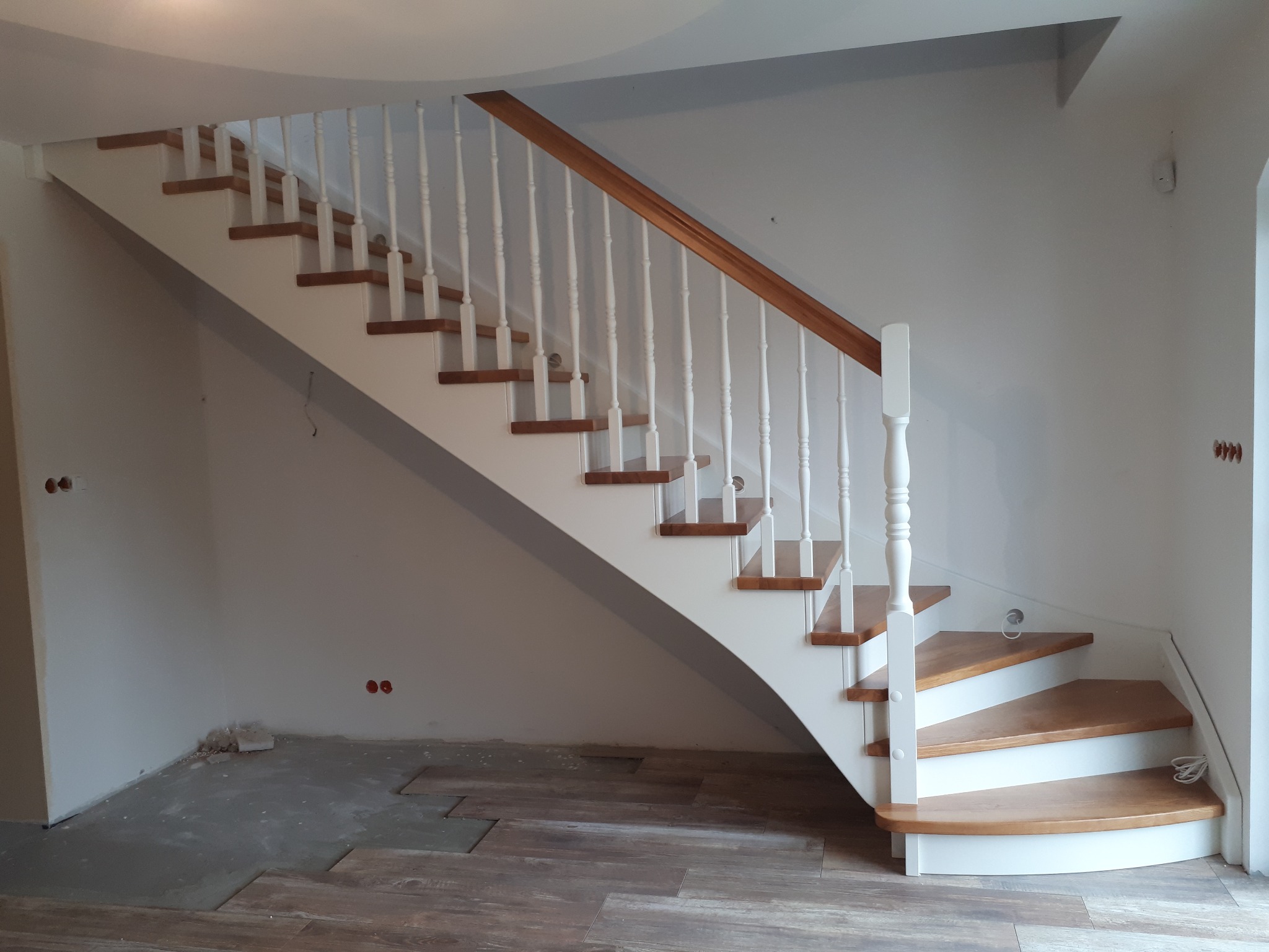 Białe schody drewniane z brązowymi stopniami i drewnianą poręczą, widok z dołu, częściowo wykończona podłoga w tle.