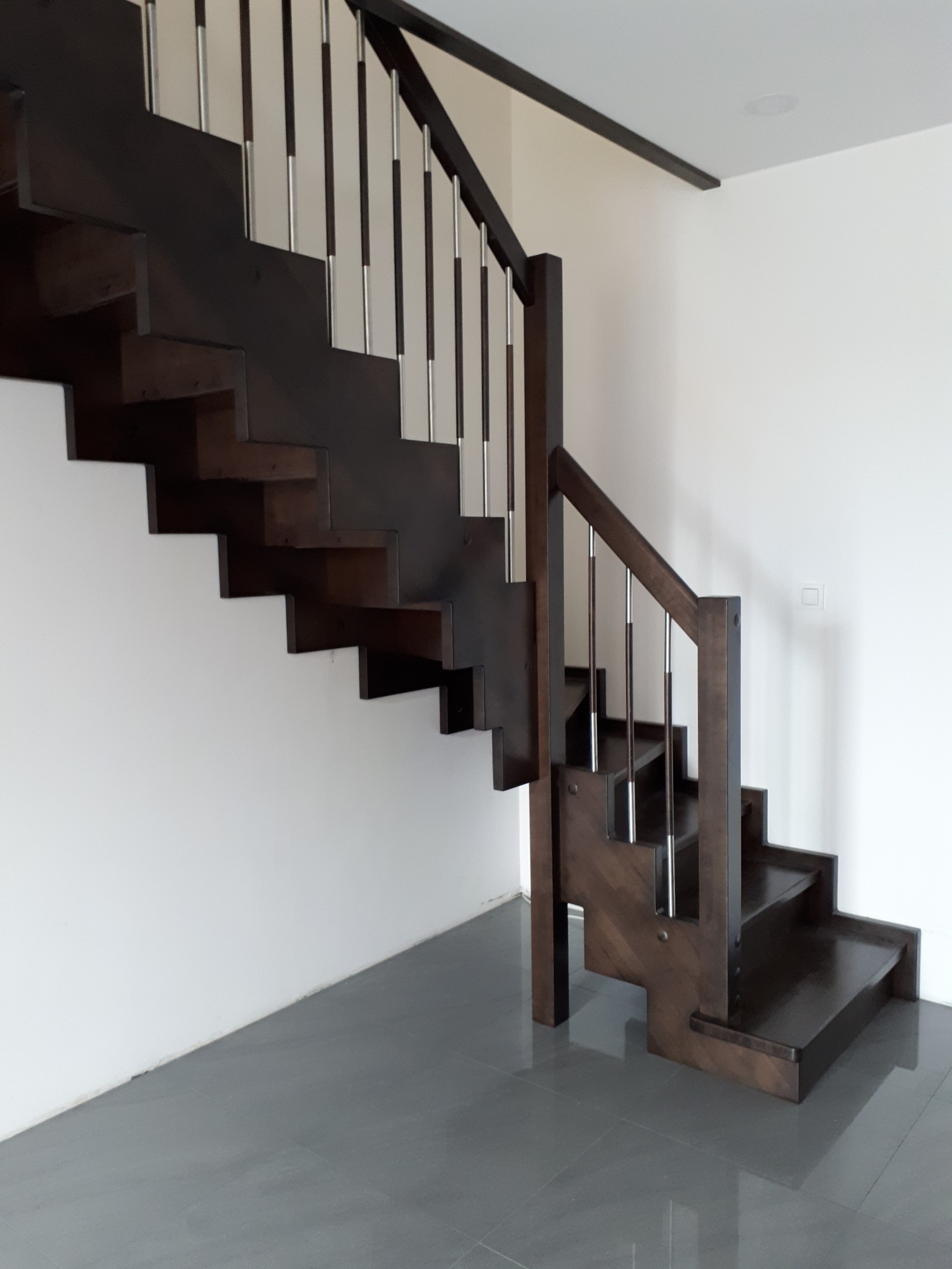 Ciemne, drewniane schody z metalowymi balustradami, w minimalistycznym wnętrzu z szarą podłogą i białą ścianą, oświetlone naturalnym światłem.