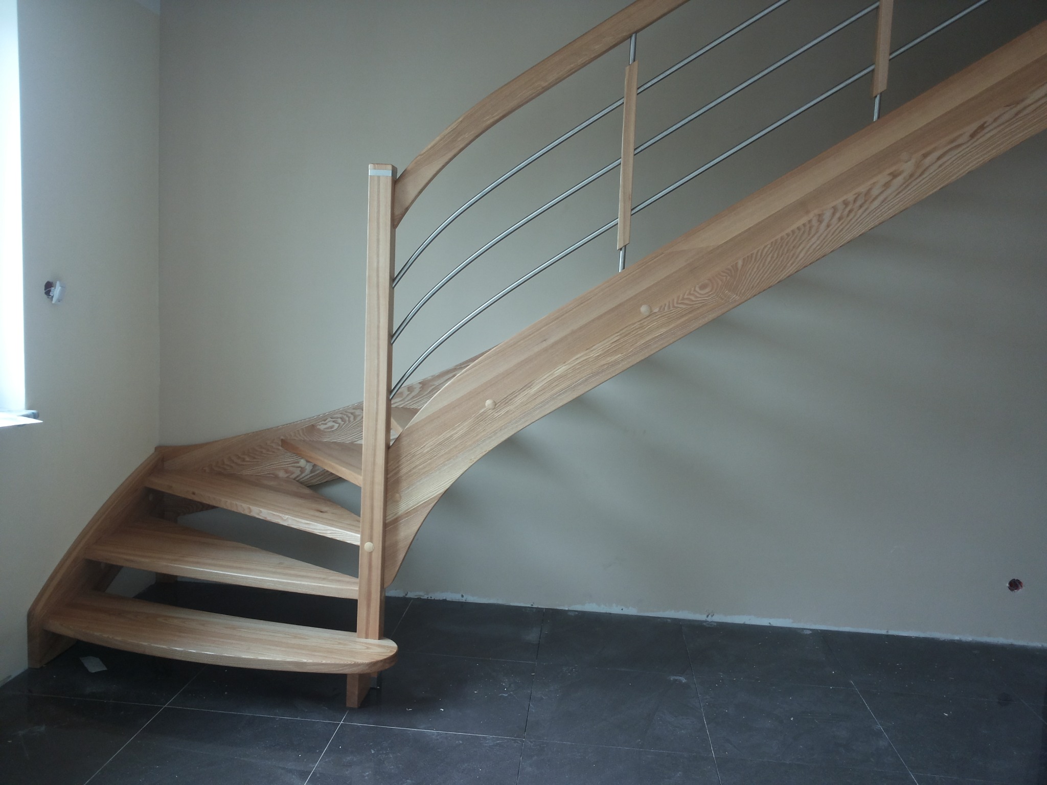 Spiralne schody z jasnego drewna z metalowymi poręczami, zamontowane w rogu pomieszczenia z szarą podłogą i beżowymi ścianami, widoczne niedociągnięcia wykończeniowe.