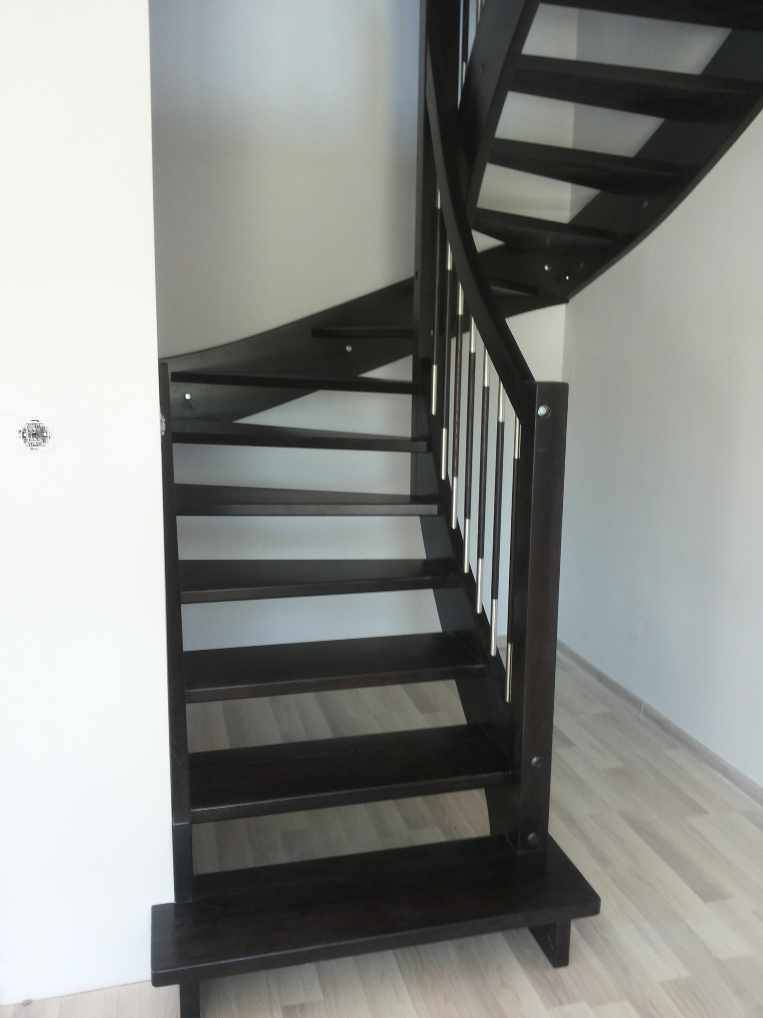 Ciemne, kręte schody z drewnianymi stopniami i metalowymi balustradami, zamontowane przy białej ścianie w jasnym wnętrzu.