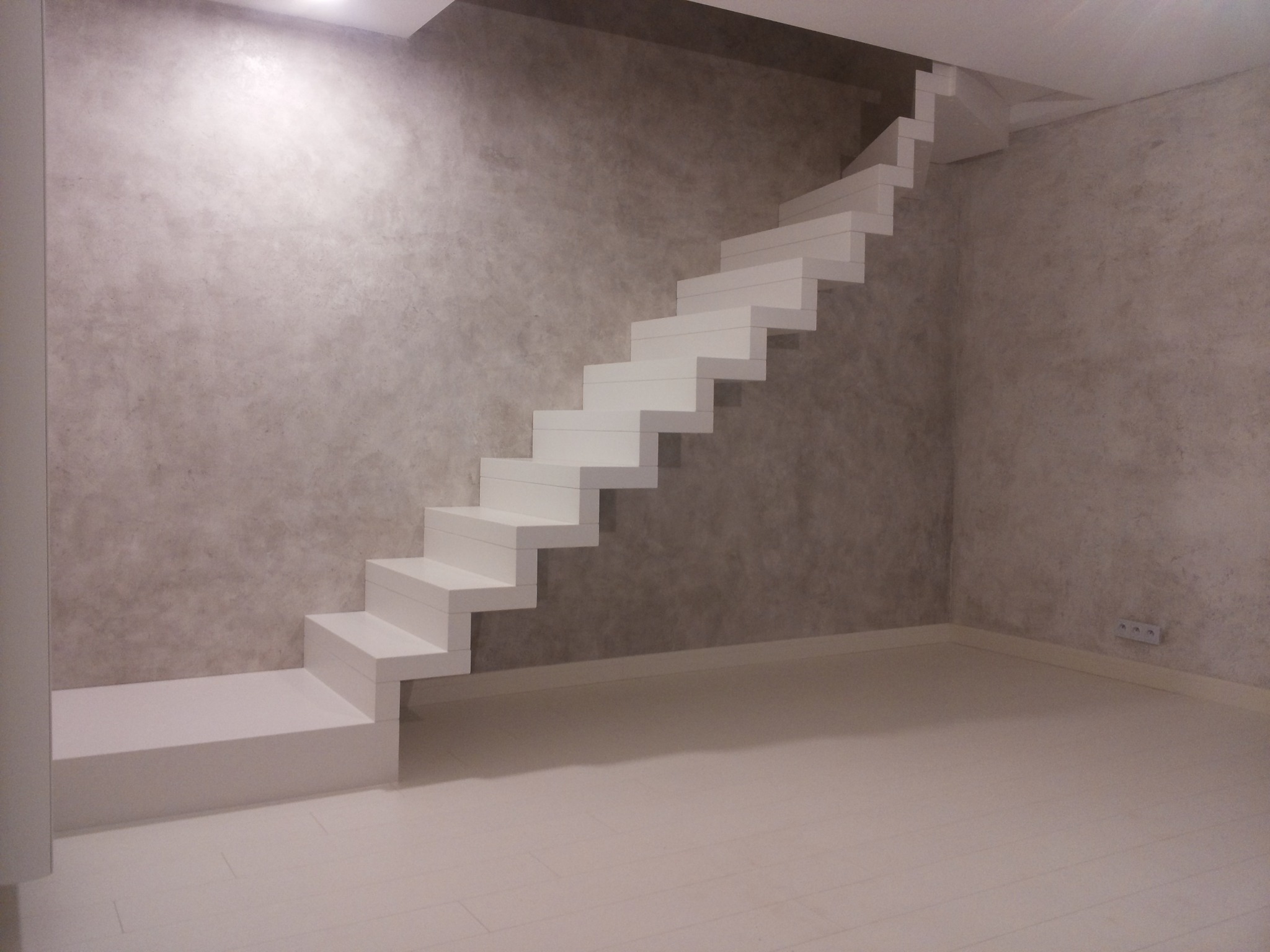 Minimalistyczne, białe schody wspornikowe na tle ściany o fakturze betonu, oświetlone od góry, widoczne gniazdka elektryczne na ścianie.