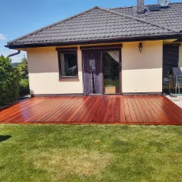 Nowoczesny, świeżo zaimpregnowany taras z desek o ciepłym, czerwono-brązowym odcieniu, przylegający bezpośrednio do domu z beżową elewacją i ciemnym dachem, otoczony zielonym trawnikiem i bujną...