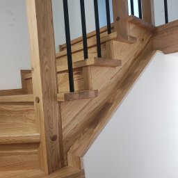 P.P.H.U. JAROSŁAW SMAGA - Drewniane schody z czarnymi, metalowymi balustradami, widoczne z dołu, z detalami wykończenia i naturalnym odcieniem drewna. Minimalistyczne tło z białej ściany.