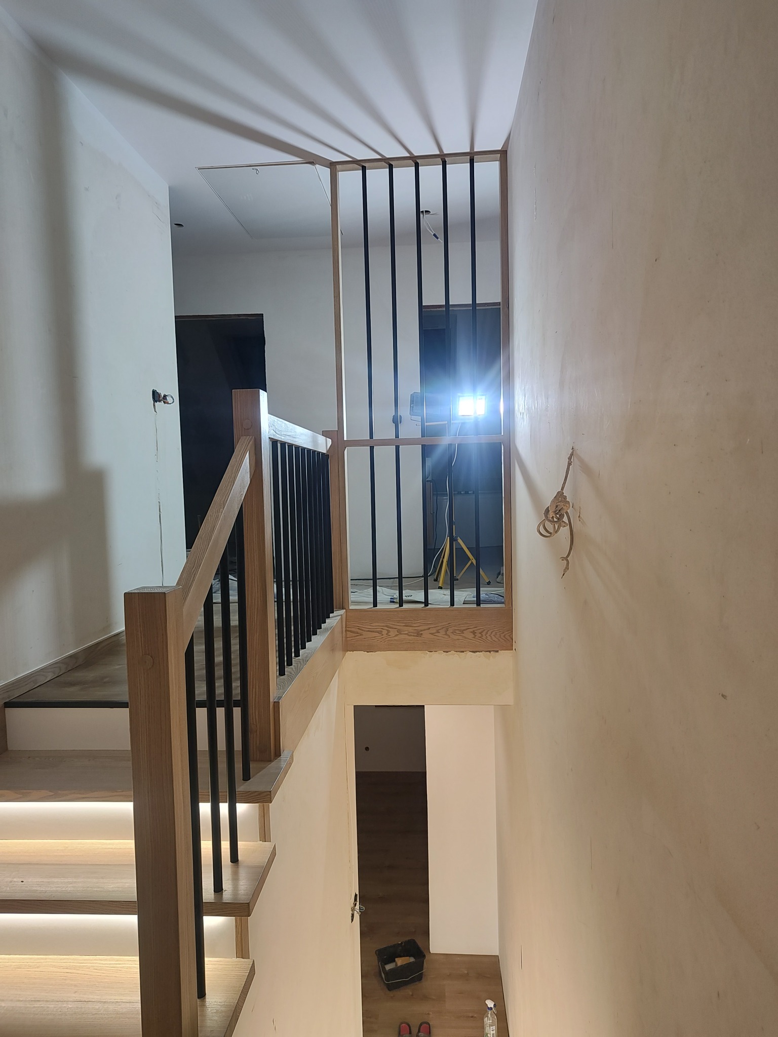 Nowoczesne schody z drewnianymi stopniami podświetlonymi LED i czarnymi balustradami. Widok z góry na dół, w tle widoczne narzędzia i lampa budowlana.