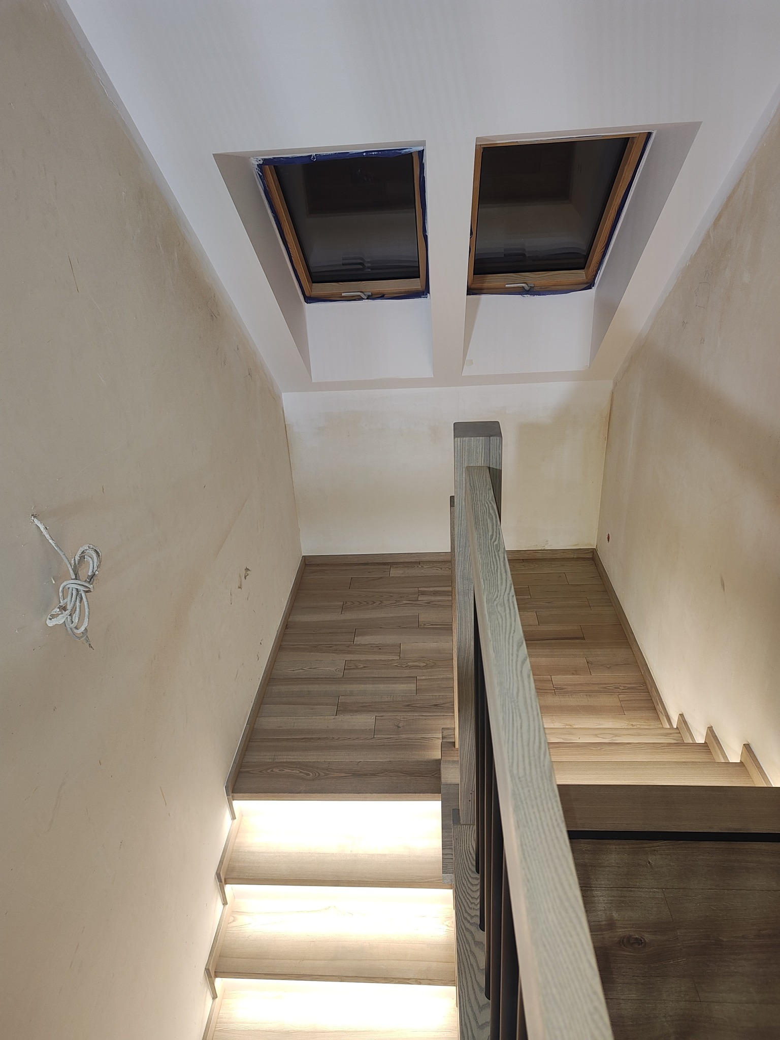 Nowoczesne schody drewniane z podświetleniem LED, widok z góry. Jasne drewno, minimalistyczny design, dwa okna dachowe w tle. Ściany w trakcie wykończenia.