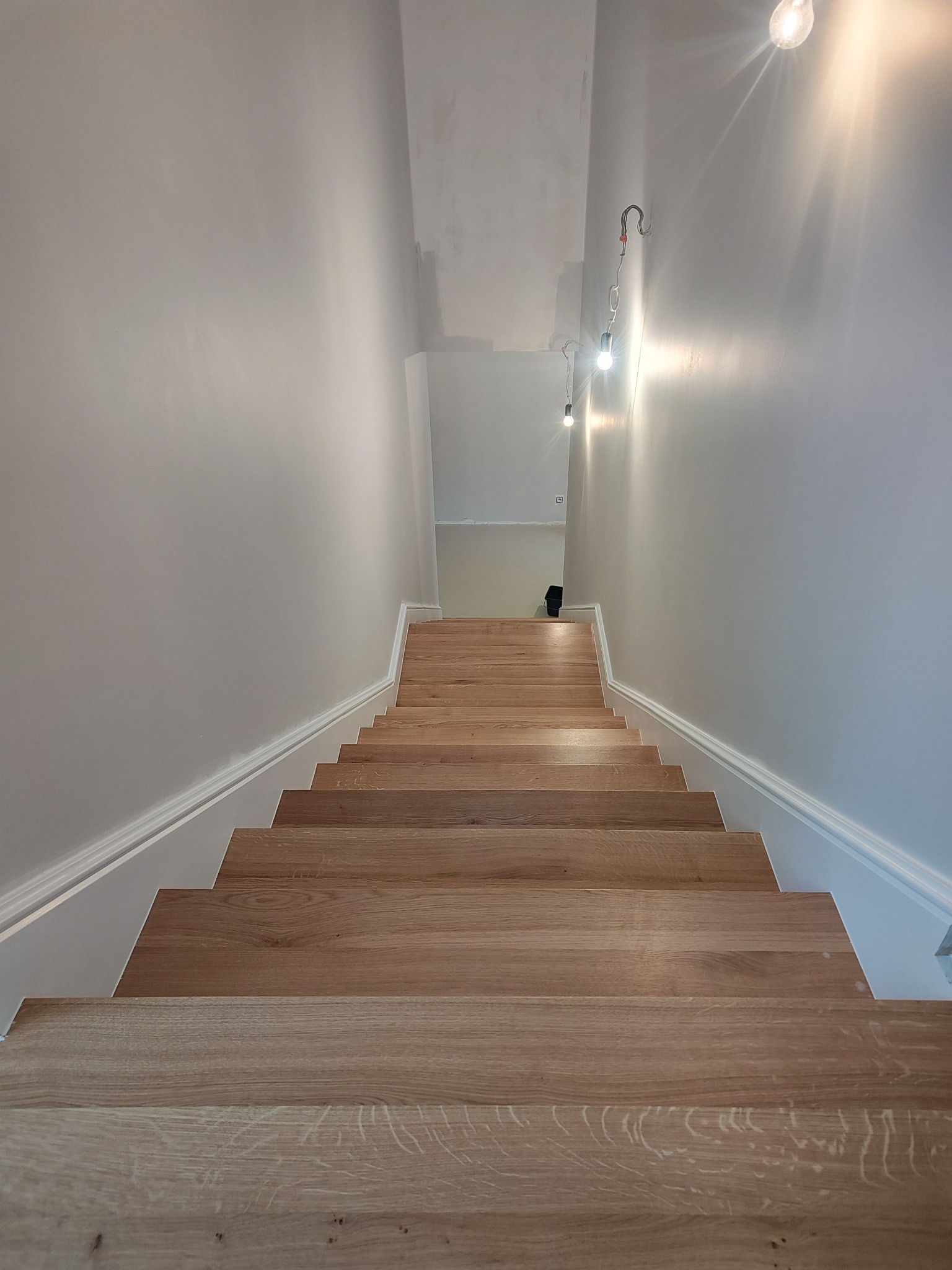 Jasne, drewniane schody z białymi listwami przypodłogowymi, widziane z góry. Na ścianie widoczne żarówki na kablach. Świeżo wykończone wnętrze.