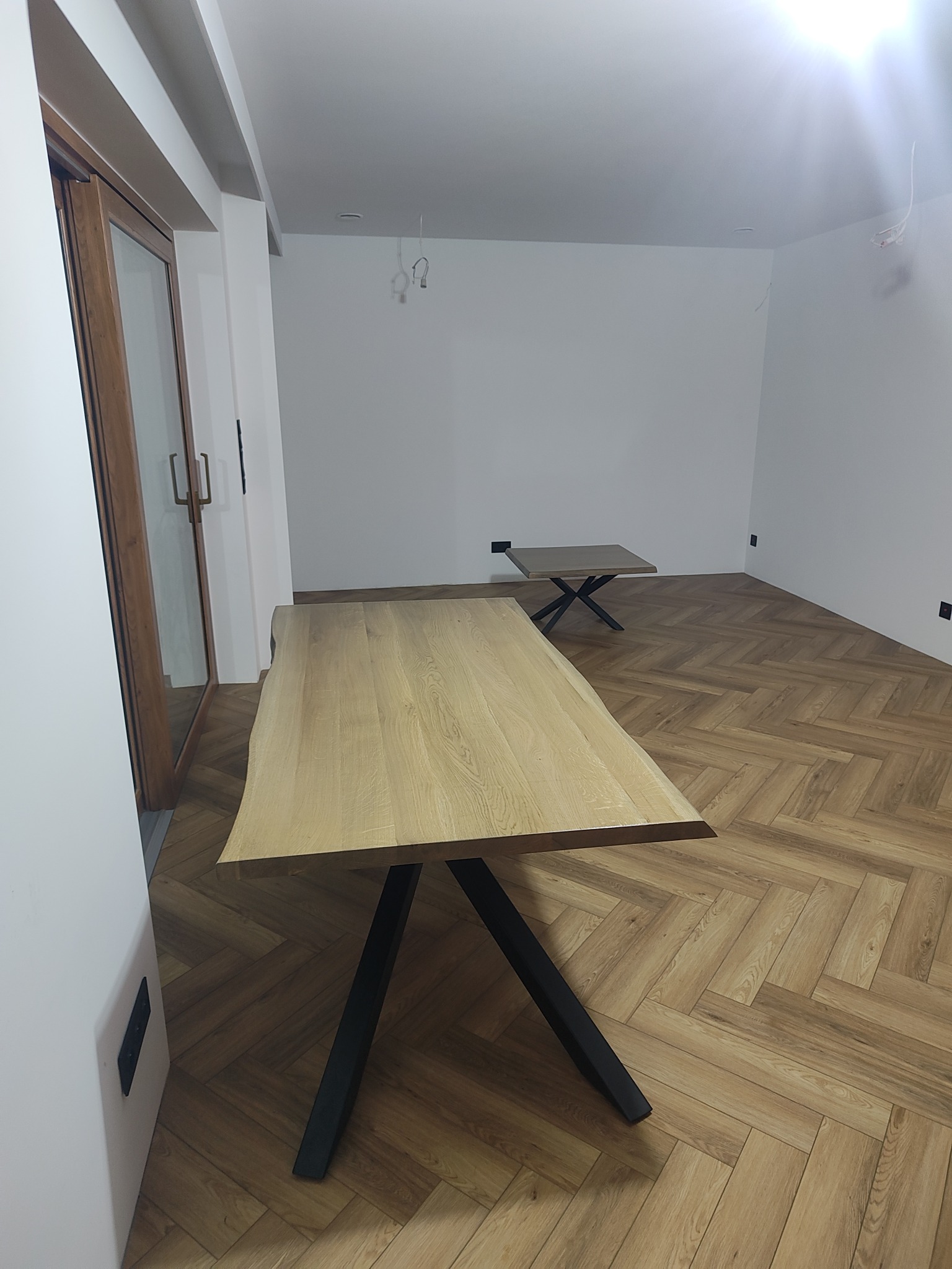 Jasne panele ułożone w jodełkę na podłodze, widoczne fragmenty dwóch stolików z drewnianymi blatami i czarnymi nogami. Minimalistyczne wnętrze z białymi ścianami.