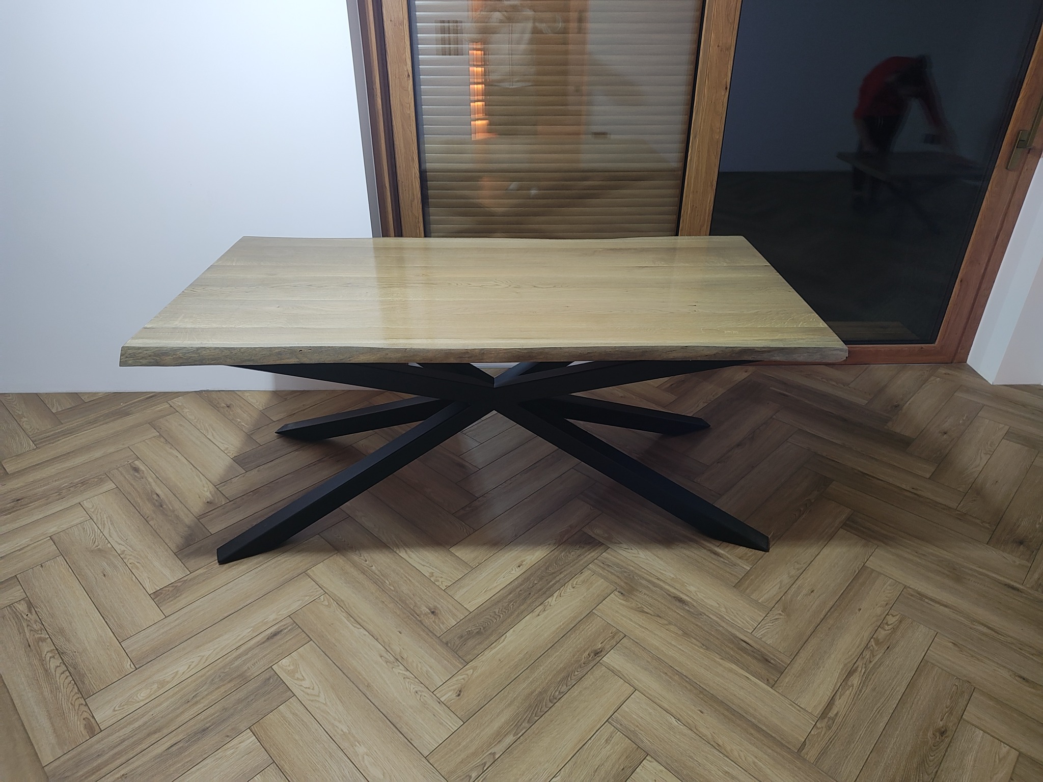 Stół z blatem z jasnego drewna o nieregularnych krawędziach i czarną, metalową podstawą w kształcie gwiazdy, na tle podłogi z paneli ułożonych w jodełkę.