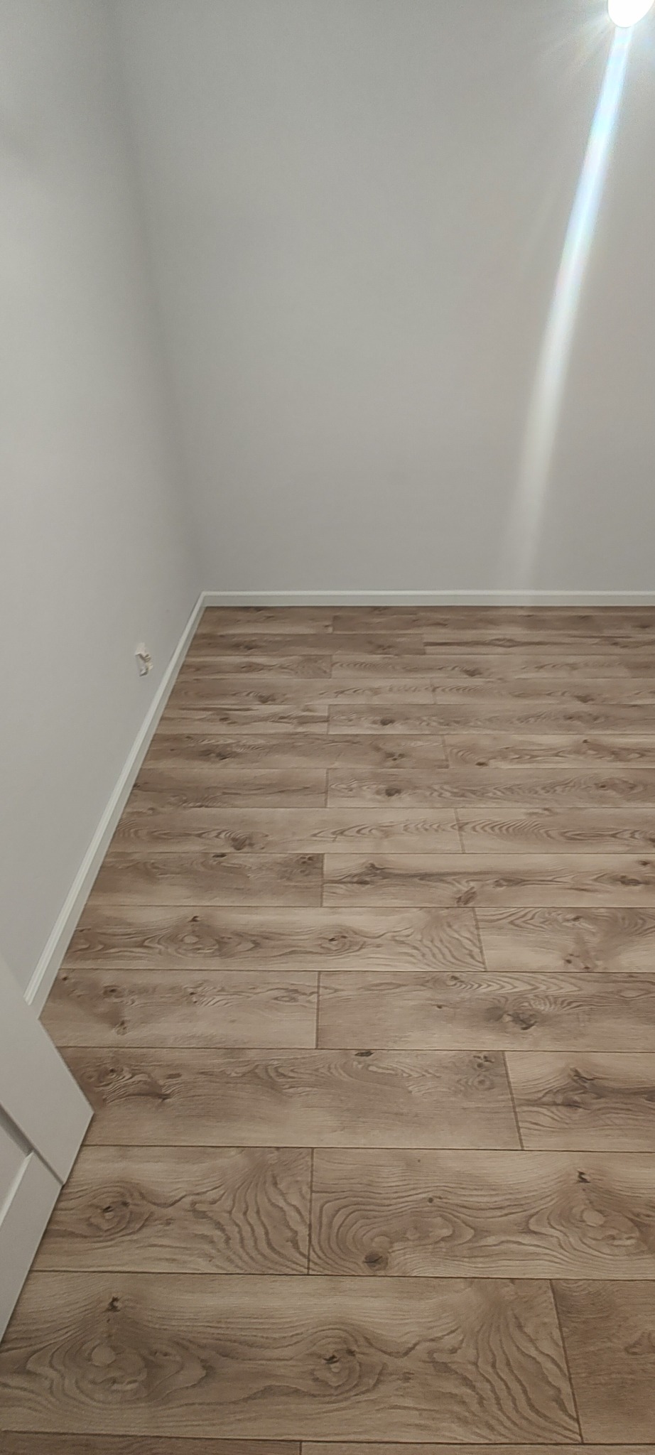 Jasne panele podłogowe z wyraźnym wzorem drewna, ułożone w nowym pomieszczeniu z białymi ścianami i listwami przypodłogowymi. Widoczny fragment drzwi.