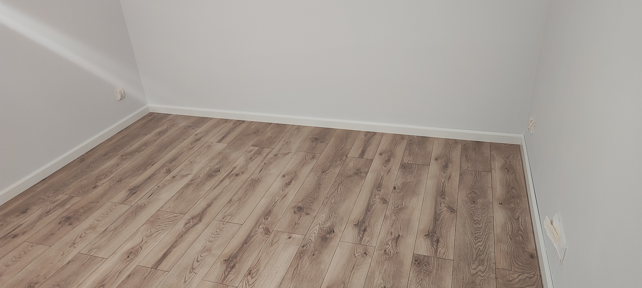 Jasne panele podłogowe ułożone w pokoju z białymi ścianami i listwami przypodłogowymi. Widoczne gniazdka elektryczne.