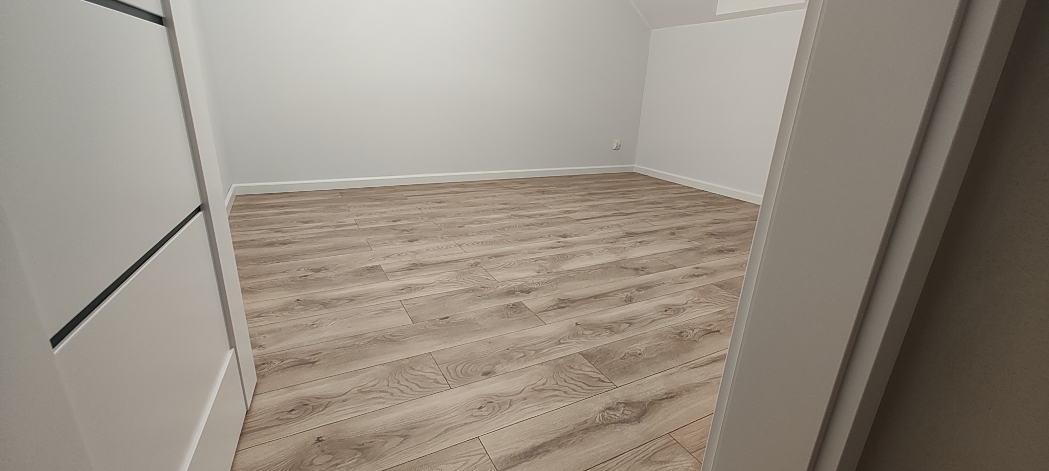 Jasne panele podłogowe z delikatnym wzorem drewna, ułożone w pustym, pomalowanym na biało pokoju. Widok zza otwartych drzwi z białą ościeżnicą i czarnym akcentem.
