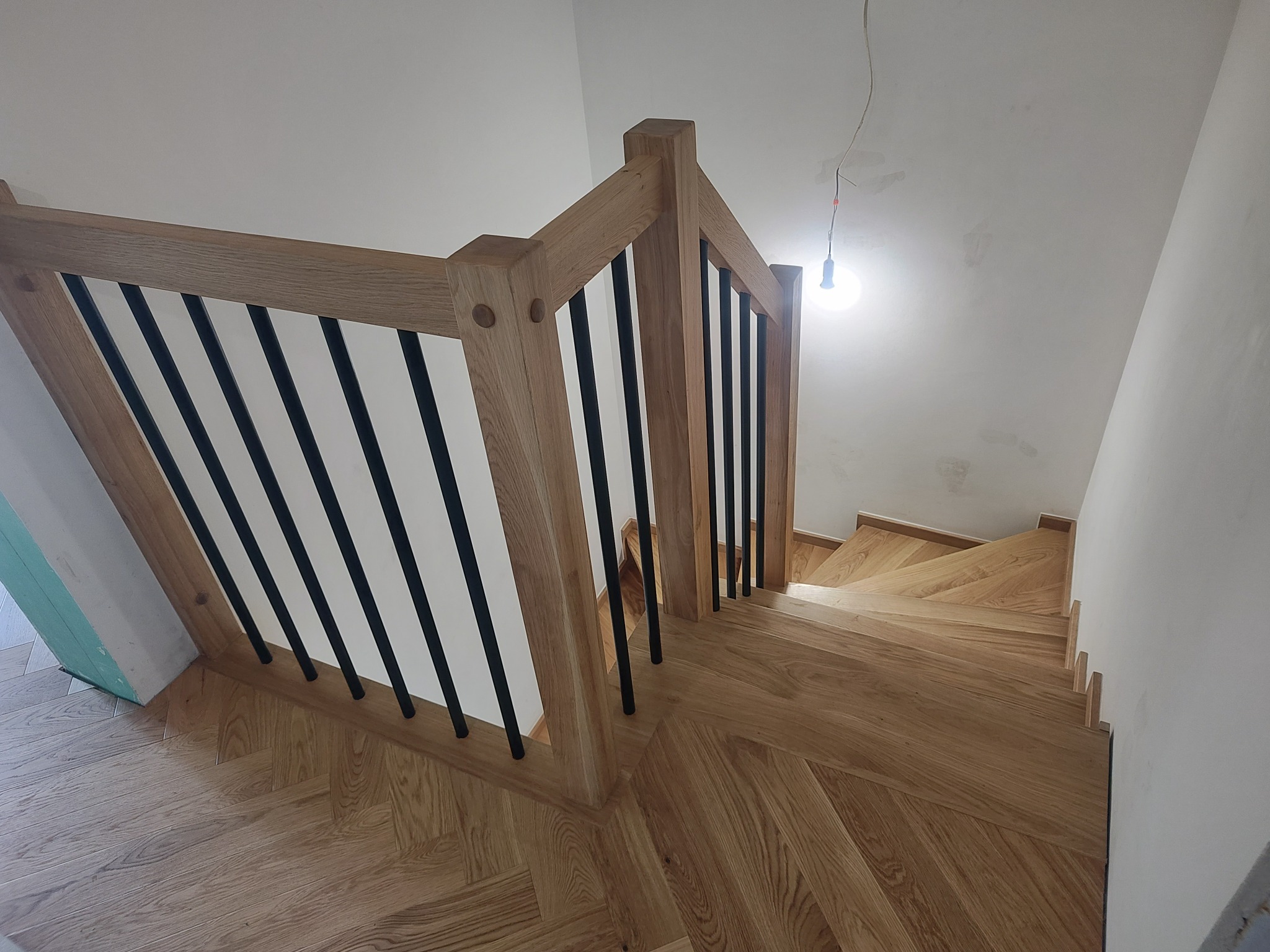 Drewniane schody z czarnymi, metalowymi balustradami, widok z góry. Parkiet ułożony w jodełkę. Surowe wykończenie ścian i wisząca żarówka.