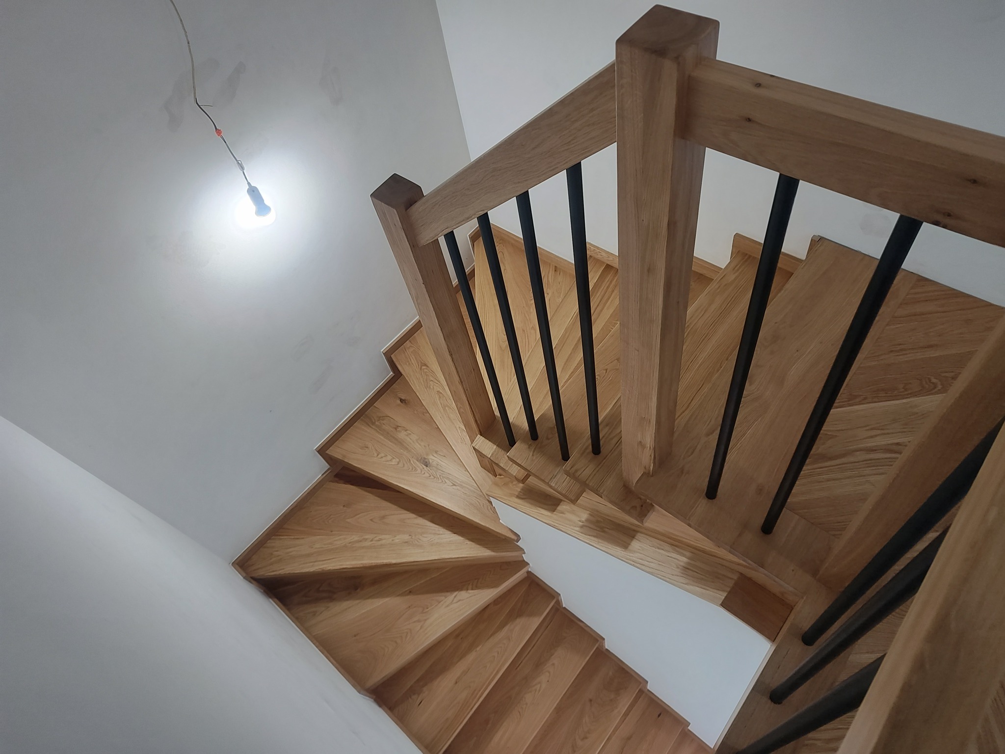 Drewniane schody z czarnymi balustradami, widok z góry. Naturalne drewno, minimalistyczny design. W tle surowa ściana z wiszącą żarówką.