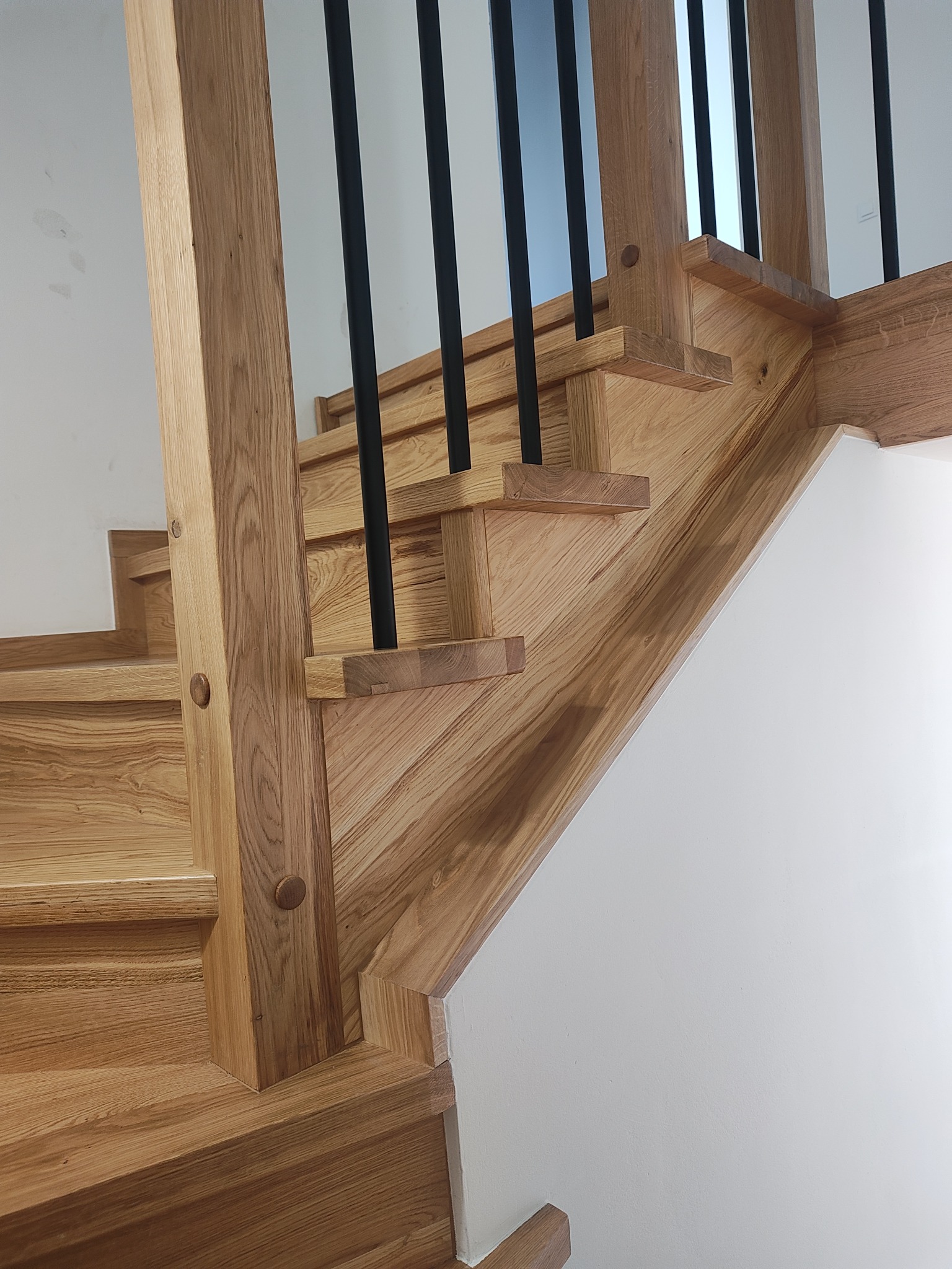 Drewniane schody z czarnymi, metalowymi balustradami, widoczne z dołu, z detalami wykończenia i naturalnym odcieniem drewna. Minimalistyczne tło z białej ściany.