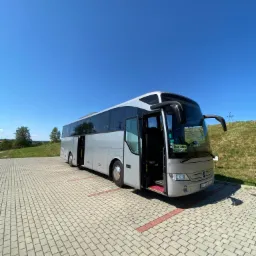 Srebrny autokar turystyczny z otwartymi drzwiami stoi na parkingu z kostki brukowej w słoneczny dzień.