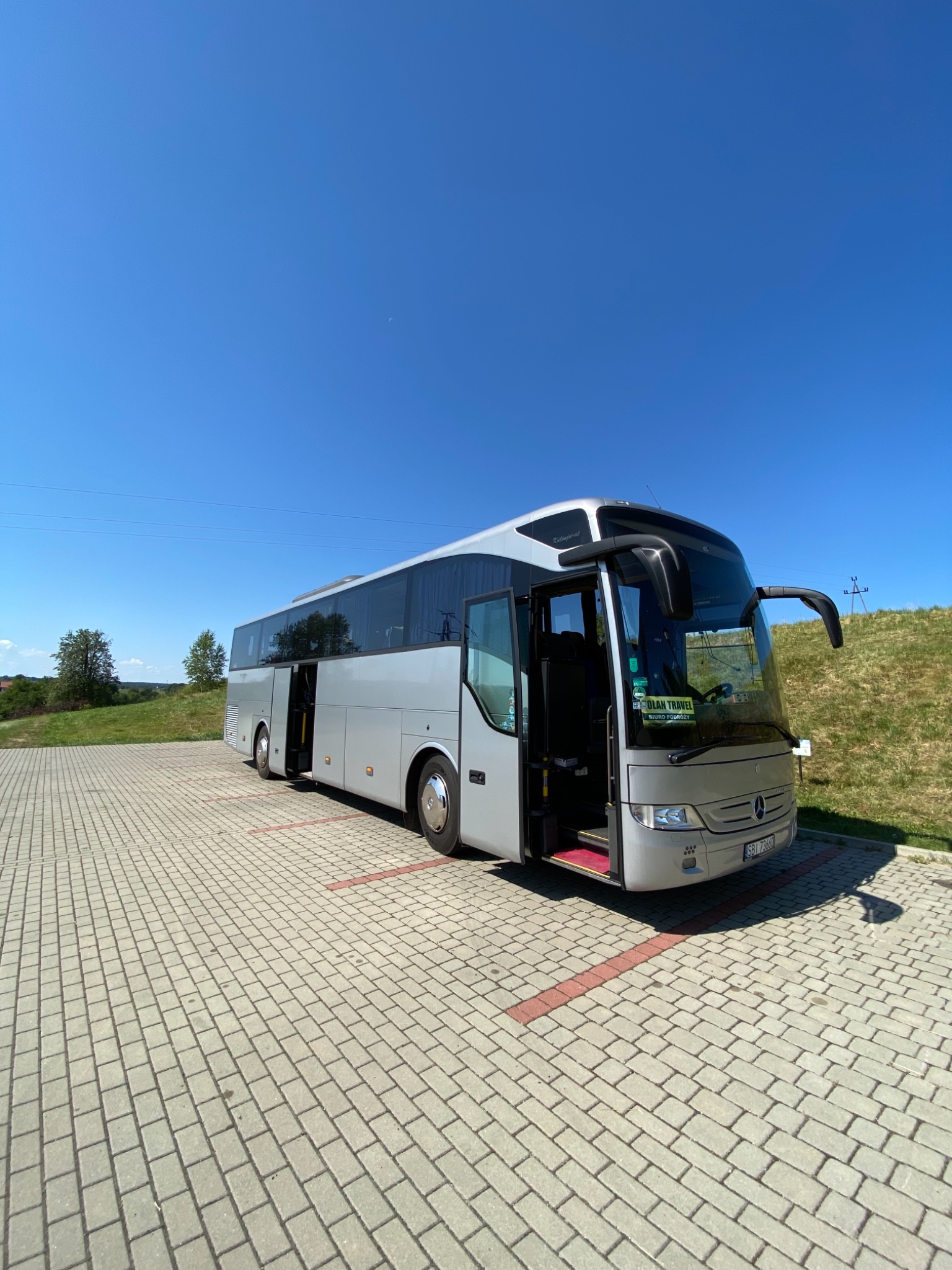Srebrny autokar turystyczny z otwartymi drzwiami stoi na parkingu z kostki brukowej w słoneczny dzień.