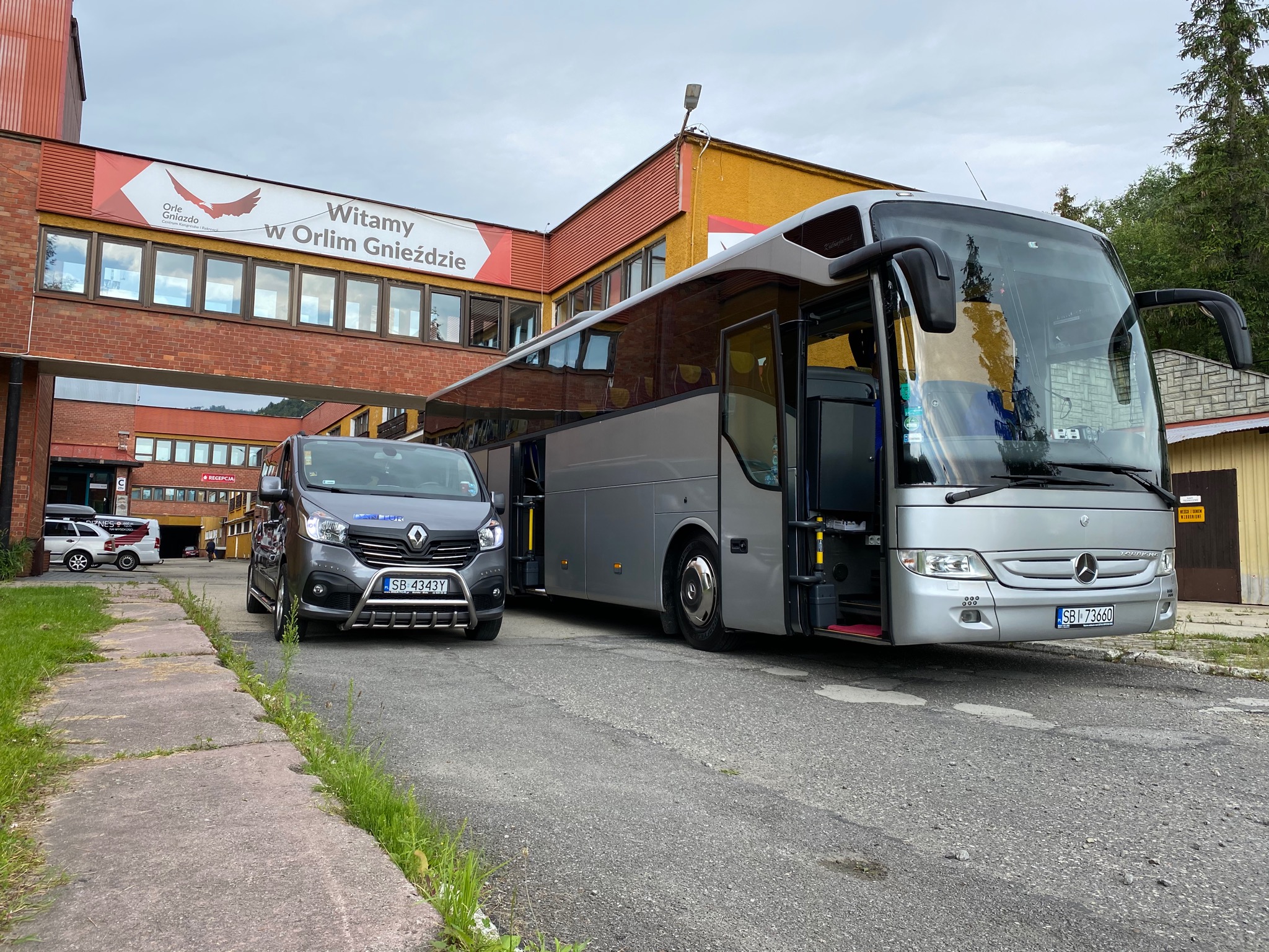 Srebrny autokar turystyczny Mercedes-Benz Tourismo i szary bus Renault Trafic na tle budynku z napisem 'Witamy w Orlim Gnieździe'.