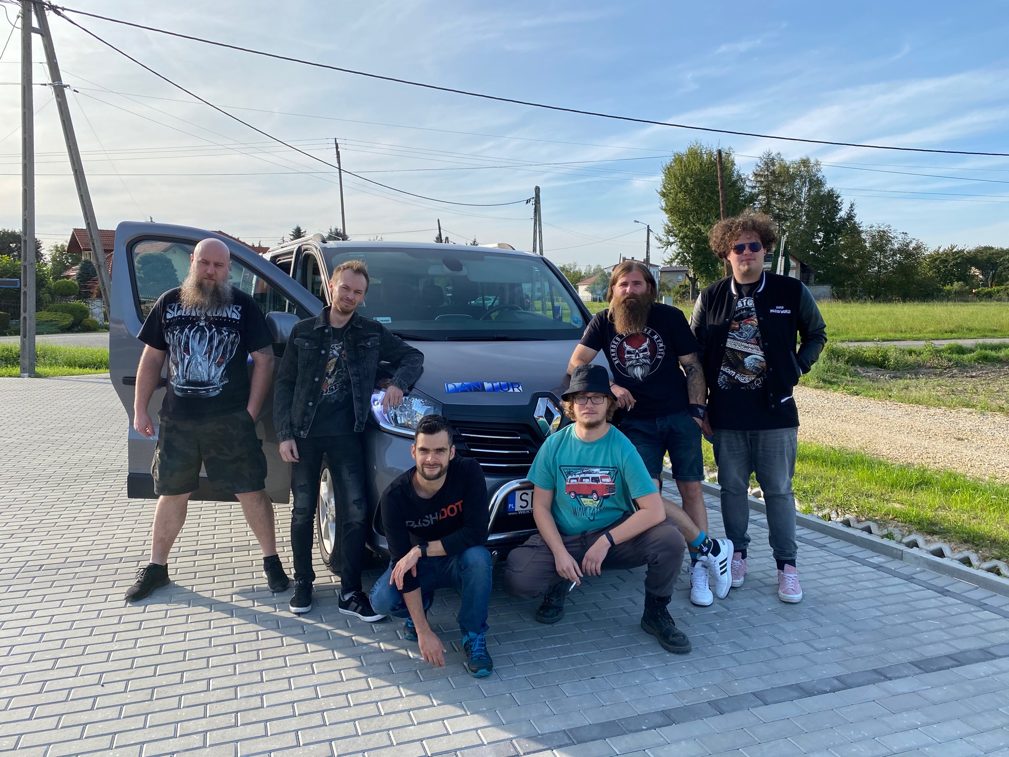 Grupa sześciu mężczyzn pozuje przy szarym vanie Renault z otwartymi drzwiami, prawdopodobnie przed wyjazdem na koncert, w tle domy i zieleń.