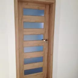 Drewniane drzwi wewnętrzne z pięcioma poziomymi przeszkleniami, widoczne na tle białych ścian i podłogi imitującej drewno.