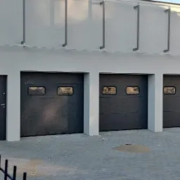 Trzy segmentowe bramy garażowe w kolorze grafitowym z prostokątnymi okienkami, obok drzwi wejściowe, widoczny system monitoringu na elewacji budynku.