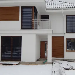 Nowoczesny dom jednorodzinny z białymi ścianami, brązowymi ramami okiennymi i drewnianymi panelami dekoracyjnymi, częściowo zasłonięty żaluzjami, zimowa sceneria.