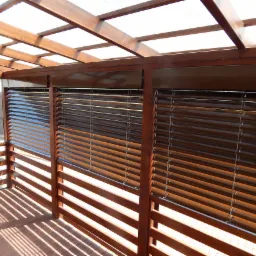 Zadaszony taras z drewnianą konstrukcją i brązowymi żaluzjami poziomymi w Kraśniku, przez które prześwieca światło.