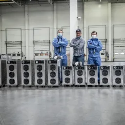 Trzech mężczyzn, dw&oacute;ch w niebieskich kombinezonach i maskach, a jeden w kraciastej koszuli, stoi za rzędem srebrnych urządzeń z wentylatorami w dużym magazynie.