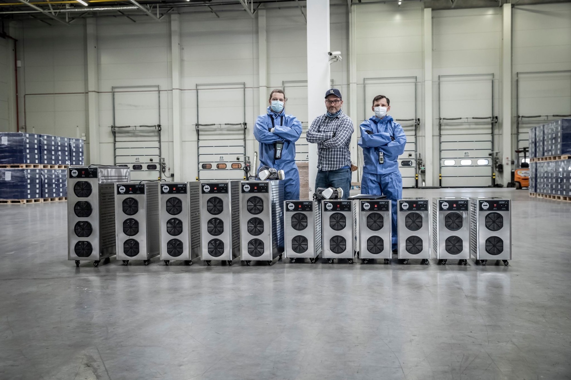 Trzech mężczyzn, dwóch w niebieskich kombinezonach i maskach, a jeden w kraciastej koszuli, stoi za rzędem srebrnych urządzeń z wentylatorami w dużym magazynie.