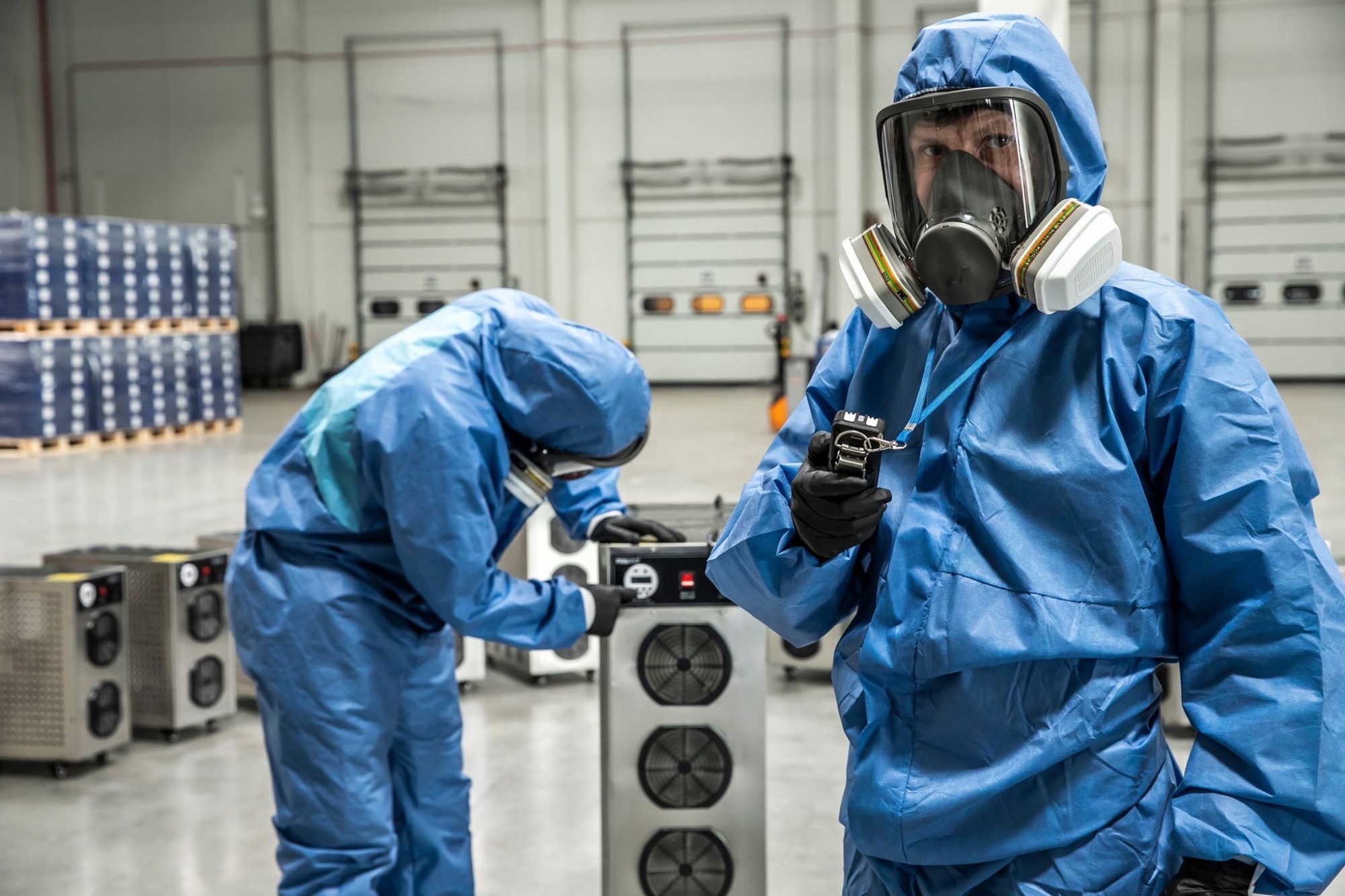 Pracownicy w kombinezonach ochronnych i maskach obsługują urządzenie filtrujące powietrze w magazynie.