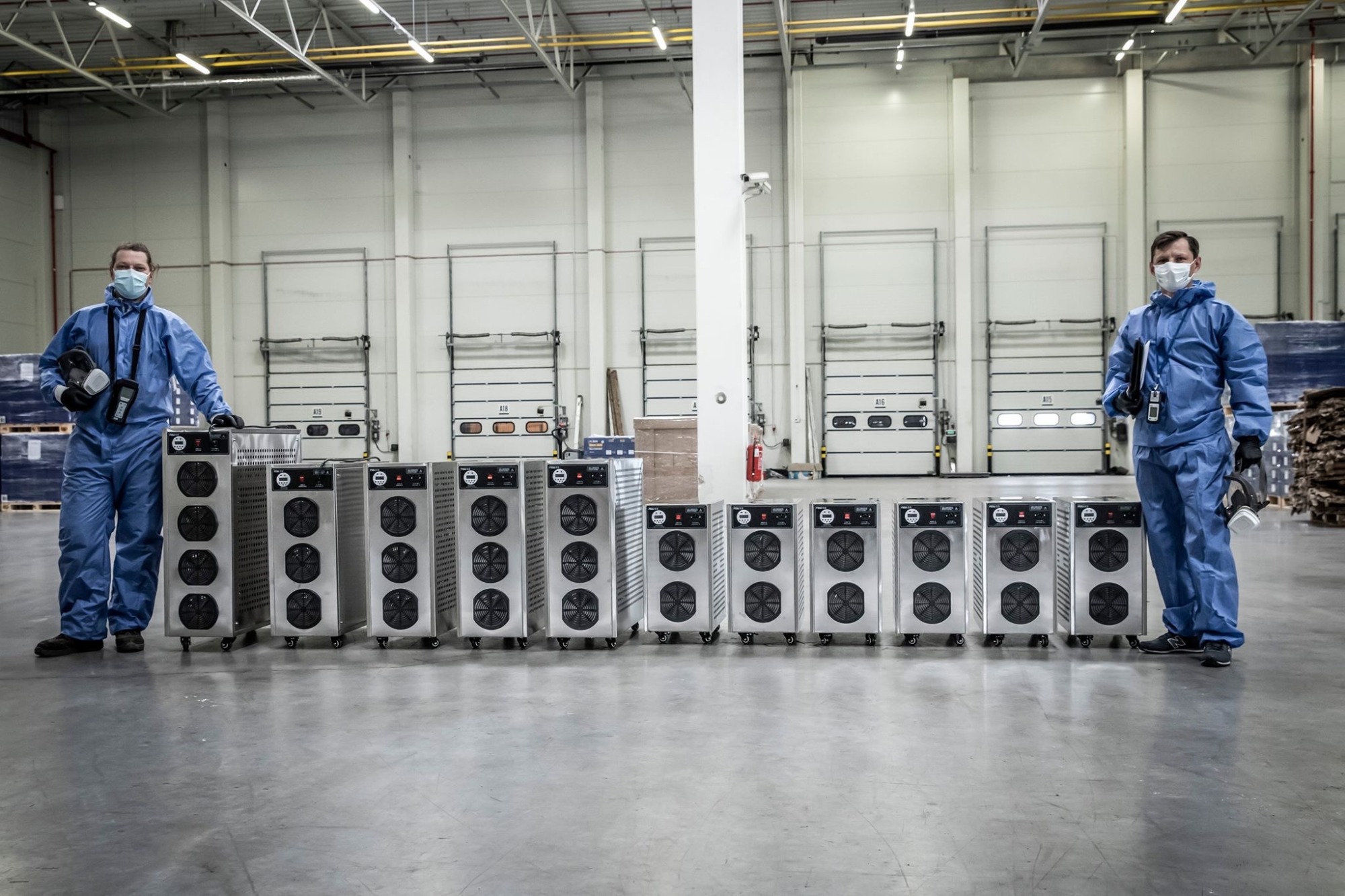 Pracownicy w kombinezonach ochronnych prezentują rząd urządzeń do dezynfekcji w hali magazynowej z bramami rolowanymi w tle.