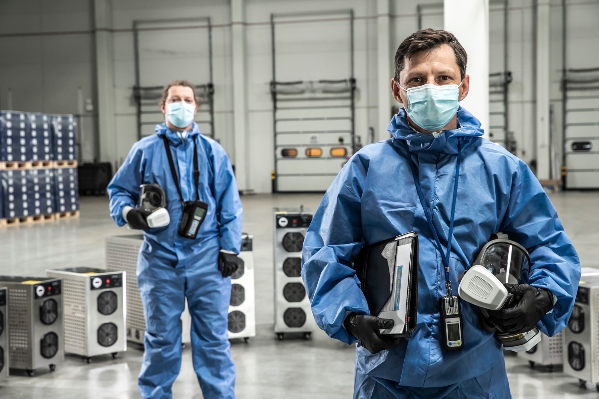 Dwóch mężczyzn w niebieskich kombinezonach ochronnych i maskach na twarzach, stojących w przestronnym magazynie obok przenośnych urządzeń filtrujących powietrze. Jeden trzyma maskę i detektor...