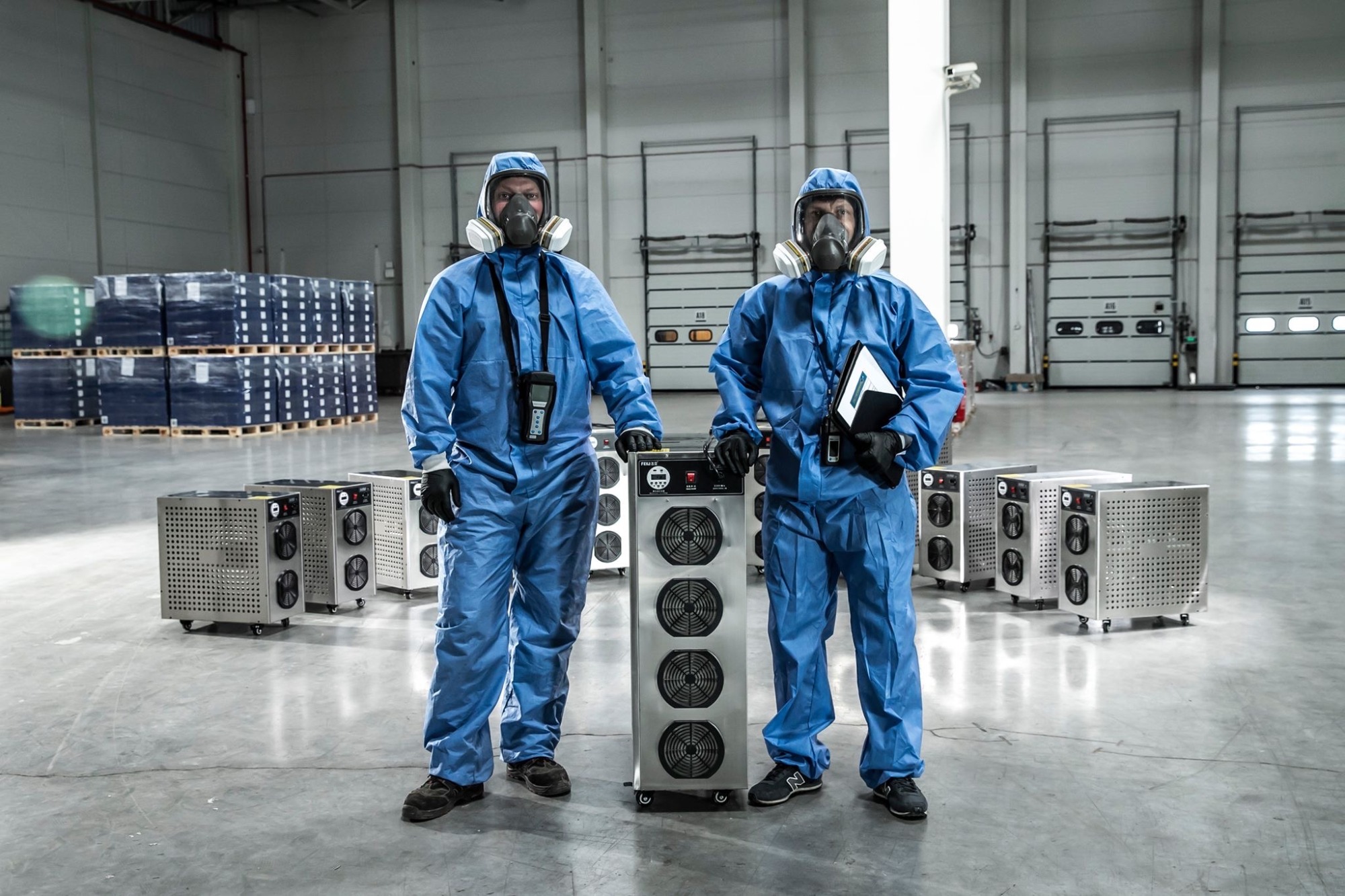 Dwóch pracowników w kombinezonach ochronnych i maskach gazowych stoi obok urządzeń z wentylatorami w hali magazynowej z paletami w tle.
